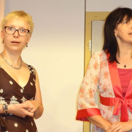 Zwei Frauen stehen nebeneinander, eine trägt eine Brille und ein braunes Kleid, die andere ein Blumenkleid mit einem roten Band.