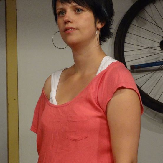 Eine Frau mit kurzen schwarzen Haaren steht vor einer weißen Wand. Sie trägt ein rosa Oberteil mit einem weißen Unterhemd und silberne Creolen. Hinter ihr ist ein Fahrradrad an der Wand befestigt.