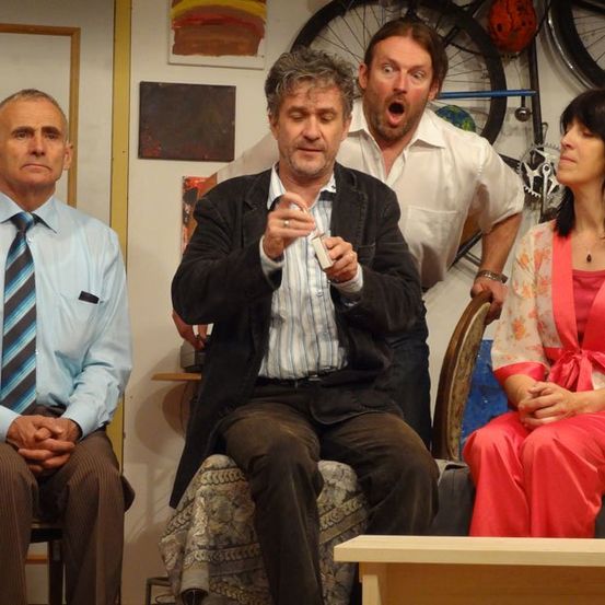 Vier Personen befinden sich in einem Raum, wahrscheinlich ein Theaterset. Drei Männer und eine Frau. Ein Mann hält eine Fernbedienung, ein anderer steht hinter ihnen, und die Frau sitzt auf einem Stuhl.