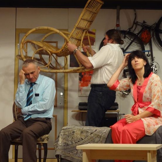 Drei Personen stehen auf einer Bühne, wobei ein Mann einen Stuhl über ihnen hebt. Ein Mann in einem blauen Hemd sitzt auf einem Stuhl, während eine Frau in einem roten Kleid auf einer Bank sitzt. Dahinter steht ein Mann in einem weißen Hemd mit einem Korb.