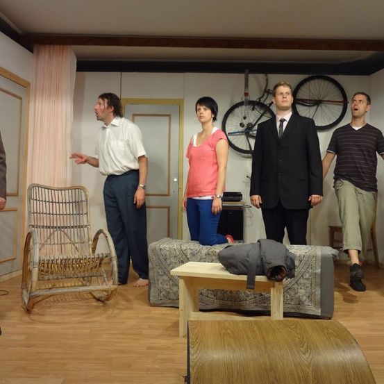 Eine Gruppe von Menschen steht in einem Raum mit Holzboden, an der Wand hängen Fahrräder und es gibt Möbel. Sie scheinen in ein Gespräch vertieft zu sein.