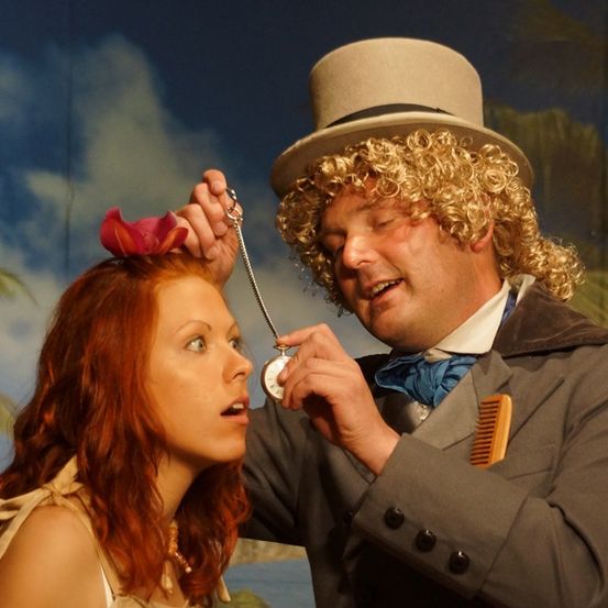 Ein Mann mit einem Tophut und einer Perücke legt einer Frau mit einer Blume im Haar eine Halskette um.