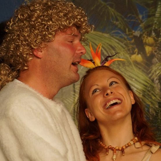 Ein Mann und eine Frau in tropischen Kostümen, der Mann mit lockigem blondem Haar und die Frau mit einem Blumenhaarschmuck, beide lächeln und schauen sich an.