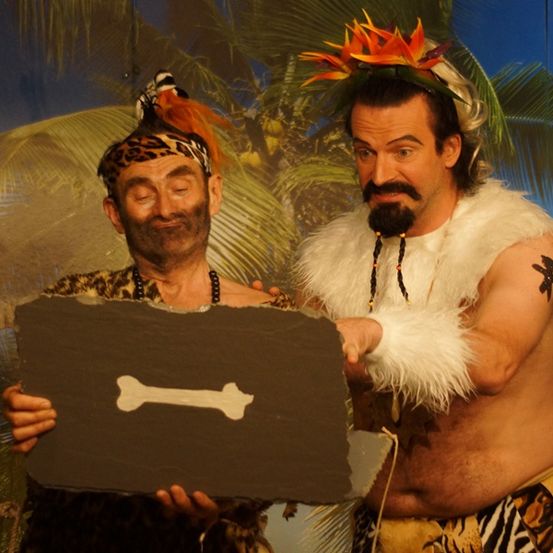 Zwei Männer, einer mit einem Leopardenmuster-Kopfband und der andere in einem Pelzoutfit, stehen vor einem tropischen Hintergrund und halten eine knochenförmige Schiefertafel.