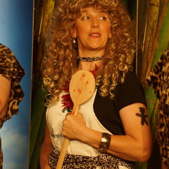 Eine Frau mit lockigem Haar und einem Leopardenmuster-Schürze hält einen Holzlöffel mit roten Flecken. Sie trägt ein schwarzes Oberteil mit einem roten und weißen Design. Der Hintergrund zeigt ein verschwommenes Leopardenmuster.