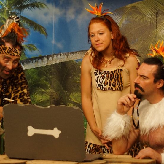 Drei Personen in Stammeskleidung, mit einer Frau in der Mitte, stehen um einen Tisch mit einem knochenförmigen Objekt.