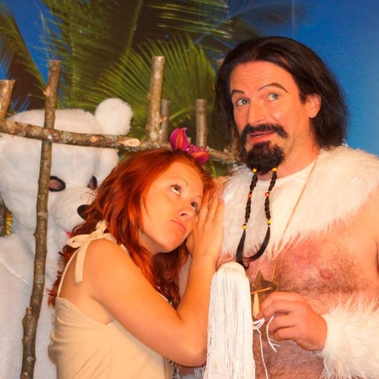 Eine Frau lehnt sich an einen Zaun und flüstert einem Mann in einem Fellschürze mit Tattoos zu. Hinter ihnen steht ein weißer Teddybär vor einer bemalten Kulisse.