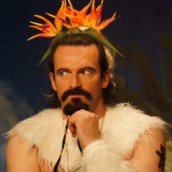 Ein Mann mit einem weißen Pelz-Vest, einem Bart und einer Blumenkrone steht vor einem blauen Hintergrund.