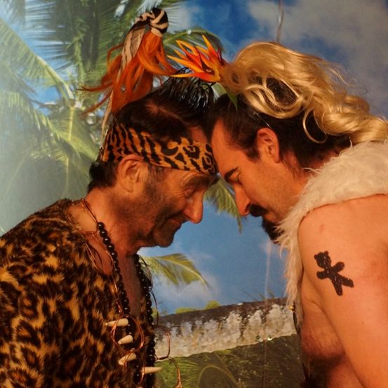 Zwei Männer in Stammeskleidung, einer mit einem Leopardenmuster-Hemd und der andere in einem weißen Pelzkostüm, stehen sich gegenüber.