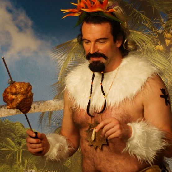 Ein Mann mit einem weißen Pelzhed und Bart hält einen Spieß mit Fleisch. Er steht vor einer Kulisse aus Palmen und blauem Himmel.