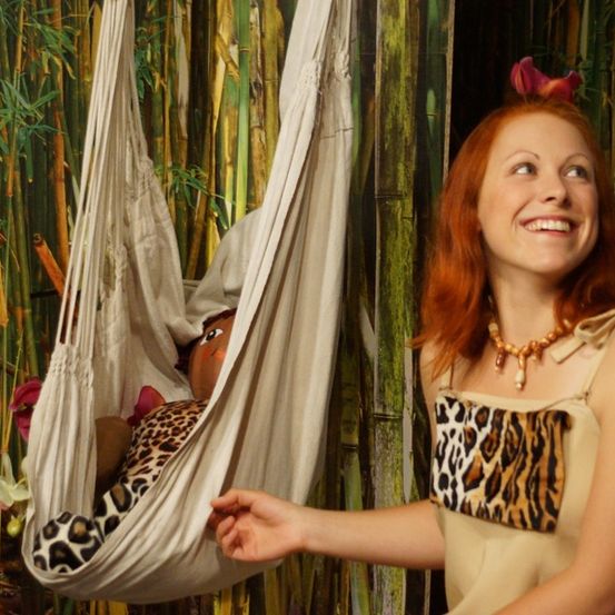 Eine lächelnde Frau in einem Leopardenmusterkleid hält eine Babypuppe in einer Hängematte und posiert vor einem Bambushintergrund.