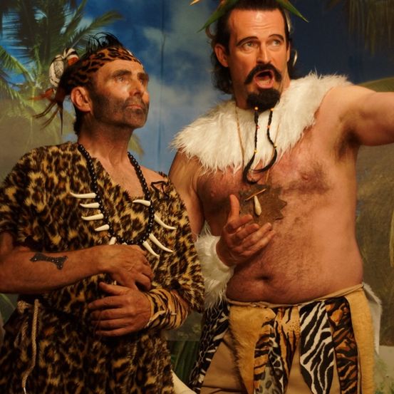 Zwei Männer in Dschungel-Kostümen stehen vor einem gemalten Hintergrund. Einer trägt einen Leoparden-Robe und eine Halskette, der andere eine Pelzkappe und eine Blume auf dem Kopf.