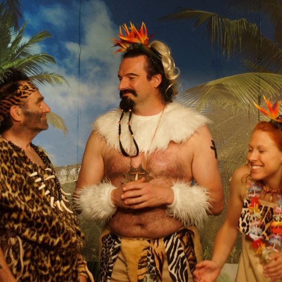 Drei Menschen in Dschungelkostümen stehen zusammen, einer in der Mitte lächelt und hält etwas in den Händen.