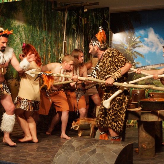 Eine Gruppe von Menschen in Tierkostümen tritt auf der Bühne auf, hält Stöcke und ein Hund ist in der Nähe.