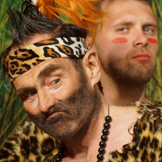 Ein Mann in einem Leoparden-Kostüm und Kopfband mit einem anderen Mann mit rotem Gesichtsmake-up dahinter.