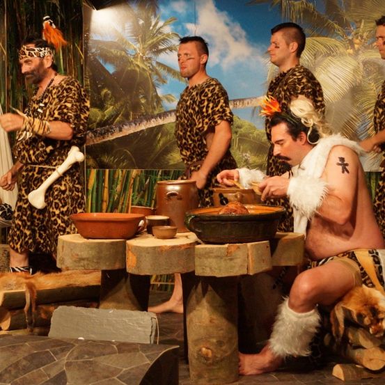 Eine Gruppe von Männern in Tierkostümen führt auf der Bühne auf, einer sitzt und bereitet Essen zu.