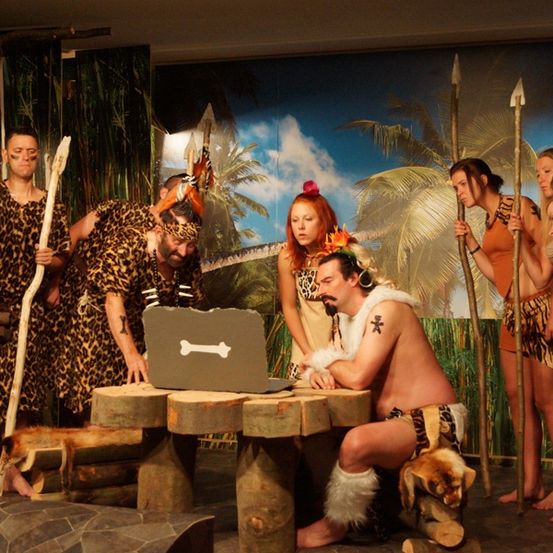 Eine Gruppe von Menschen in Leopardendruck-Kostümen steht auf der Bühne, einer kniet mit einem Laptop. Hinter ihnen halten andere Speere. Ein Hintergrund zeigt Palmen.