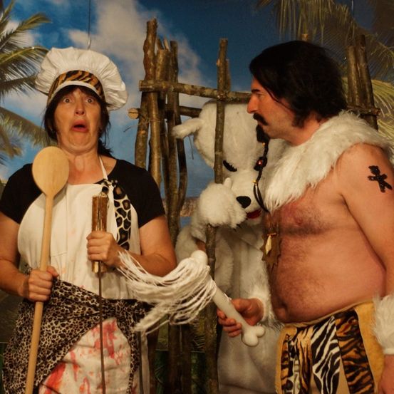 Eine Frau mit einem Holzlöffel steht neben einem Mann in einem Leopardenkostüm, mit einem weißen Hund in einem Pelzkragen hinter ihnen, vor einem tropischen Hintergrund posierend.