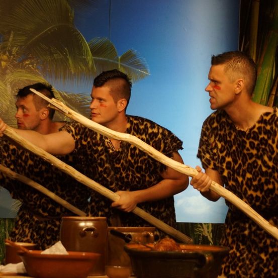 Drei Männer in Leopardenmuster-Kleidung halten Holzstäbe. Sie stehen offenbar auf einer Bühne, mit einer Kulisse, die tropische Szenerie zeigt.