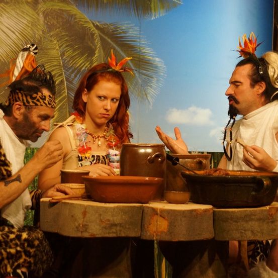 Drei Personen in Stammeskleidung sitzen um einen Tisch mit Schalen und Töpfen herum. Der Mann rechts spricht mit der Frau in der Mitte.