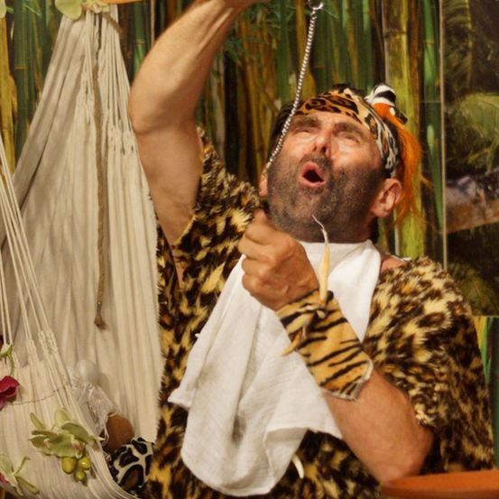 Ein Mann in einem Leoparden-Print-Kostüm führt einen Zaubertrick mit einem Holzlöffel durch, der an einer Kette hängt.