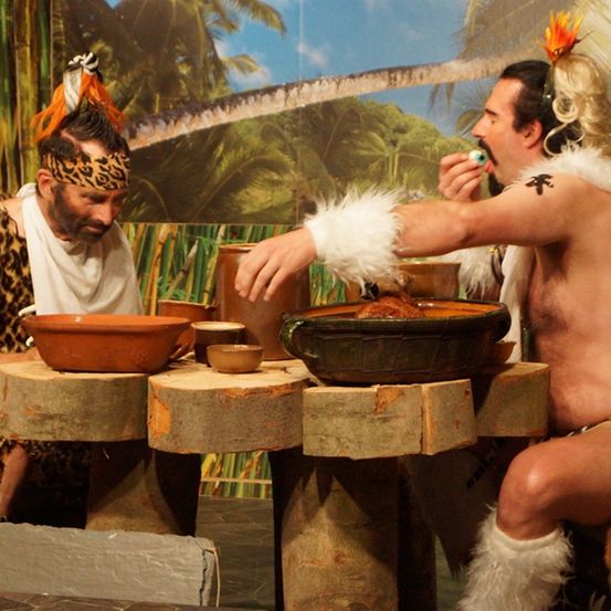 Zwei Männer in Stammeskleidung sitzen um einen Holztisch herum, einer greift nach einer Schüssel. Ein Mann hat ein Leopardenmuster um den Kopf.