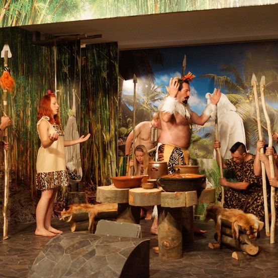 Eine Gruppe von Menschen in Leopardendruck-Kostümen führt eine Zeremonie auf einer Bühne mit Bambushintergrund durch. Ein Mann mit Kopfschmuck und Speer steht vor einem Tisch mit Schüsseln.