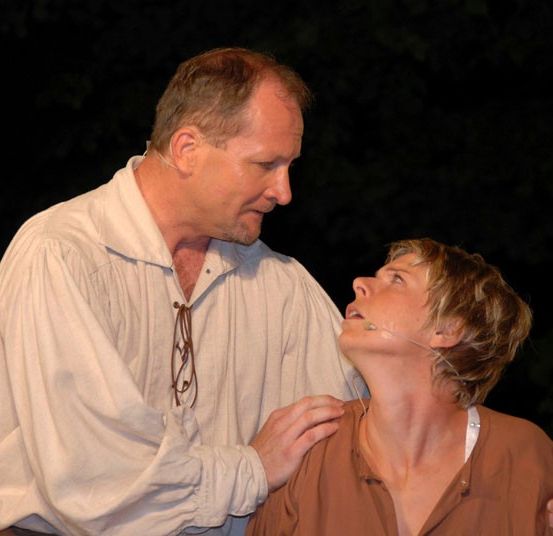 Ein Mann und eine Frau sind auf der Bühne, wahrscheinlich bei einer Aufführung. Der Mann hat eine Brille an seiner Kleidung hängen.