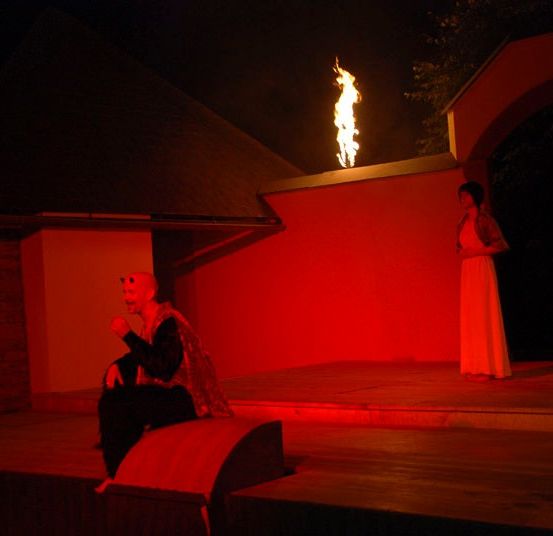 Eine nächtliche Aufführung mit einem Feuer, einem sitzenden Mann und zwei Frauen in weißen Kleidern, die unter einem rot beleuchteten Bogen stehen.