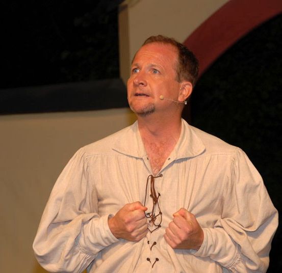 Ein Mann mit Mikrofon und Brille spricht auf einer Bühne, gekleidet in einem weißen Hemd mit langen Ärmeln.