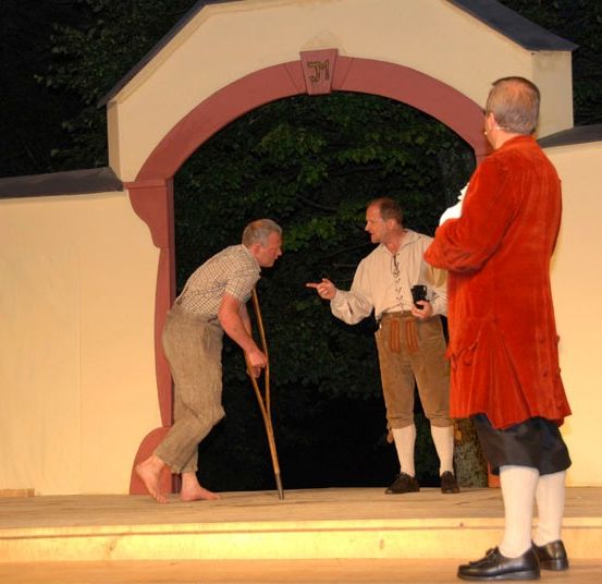 Drei Männer stehen auf einer Bühne, einer mit einem Stock, ein anderer gestikuliert. Ein Bogen und Grünpflanzen sind im Hintergrund.