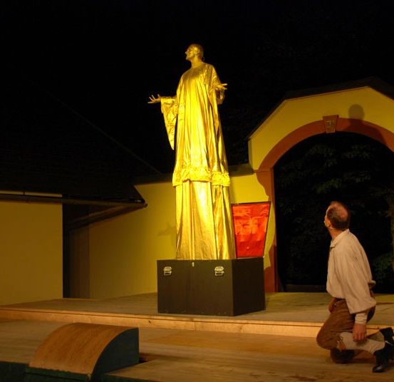 Ein Mann kniet vor einer goldenen Statue in einem schwach beleuchteten Bereich, möglicherweise bei Nacht. Die Statue steht auf einem Podest vor einem gelben Gebäude.