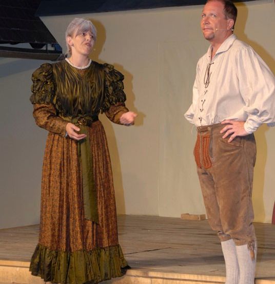 Eine Frau in historischer Kleidung steht auf der Bühne und spricht einen Mann in ähnlicher Tracht an. Beide schauen direkt in die Kamera.