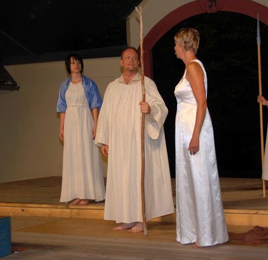 Vier Menschen in weißen Roben stehen auf einer hölzernen Bühne, jeder hält einen Stab. Der Mann in der Mitte trägt einen weißen Umhang, während die Frau links einen blauen Umhang hat. Der Hintergrund ist dunkel.