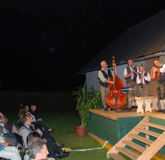 Eine Gruppe von Musikern tritt bei Nacht auf einer Bühne auf, vor einem sitzenden Publikum. Ein Mann spielt Cello, während andere Flöte und Akkordeon spielen.