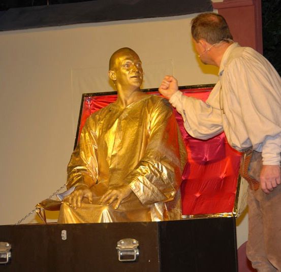 Ein Mann mit Mikrofon steht neben einer goldenen Statue in einer roten Kiste, möglicherweise auf der Bühne.