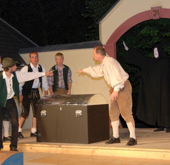 Eine Gruppe von Menschen auf einer Bühne mit einer Holztruhe und einer Person in einem schwarzen Gewand.