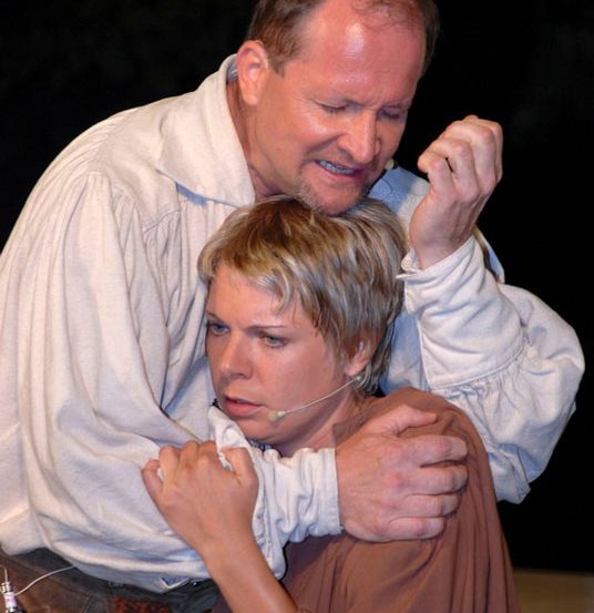 Ein Mann in einem weißen Hemd und eine Frau in einem braunen Kleid führen auf der Bühne eine Szene auf. Der Mann hält die Frau mit seinen Armen fest.