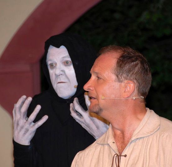 Ein Mann in einem weißen Hemd steht einem Mann in einem schwarzen Umhang und Maske gegenüber, wahrscheinlich bei einer Aufführung.