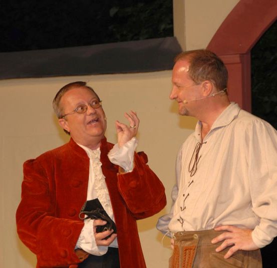 Zwei Männer stehen, einer in einem roten Mantel, der ein kleines Objekt hält, der andere in einem weißen Hemd mit Mikrofon, beide lächeln und führen ein Gespräch.