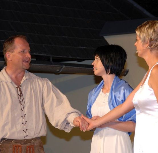 Drei Personen stehen auf einer Bühne, wobei der Mann in der Mitte die Hände von zwei Frauen hält. Der Mann trägt ein weißes Hemd mit Schnürsenkeln und einem Gürtel. Die Frauen tragen ein weißes Kleid und einen blauen Umhang.