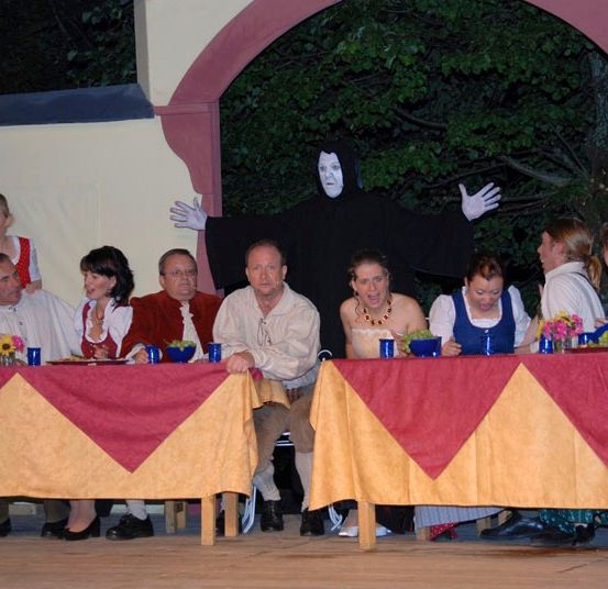 Eine Gruppe von Menschen sitzt an Tischen auf einer Bühne, mit einer Person in einem schwarzen Kostüm hinter ihnen. Die Tische sind mit rotem und gelbem Tuch bedeckt.