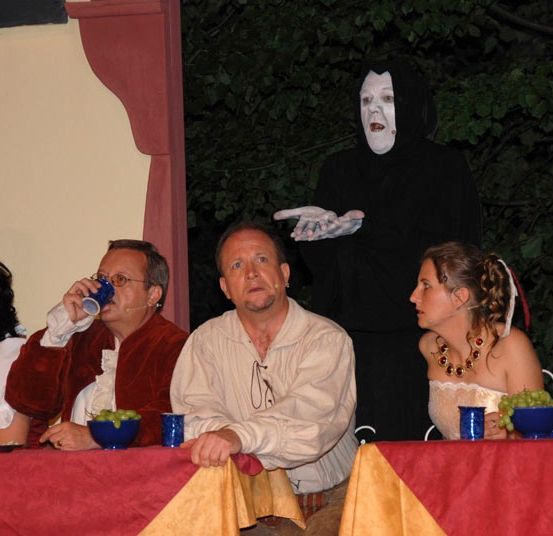 Mehrere Menschen sitzen an einem Tisch, ein Mann in einem schwarzen Kostüm steht hinter ihnen. Ein Mann trinkt aus einem blauen Becher.