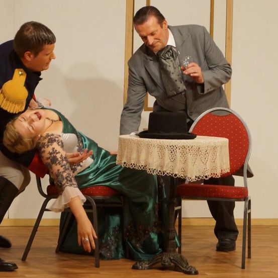 Drei Personen auf einer Bühne, eine Frau in einem grünen Kleid liegt in einem Stuhl, während ein Mann mit einer gelben Quaste über sie gebeugt ist. Ein anderer Mann hält ein Glas und einen Hut auf einem Tisch.