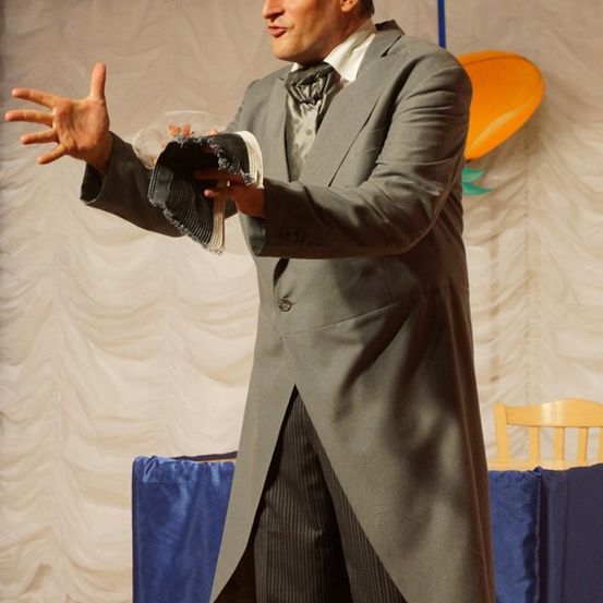 Ein Mann in einem formellen Anzug gestikuliert mit den Händen, hält einen Hut in der Hand und steht vor einem mit einem blauen Tuch bedeckten Tisch.