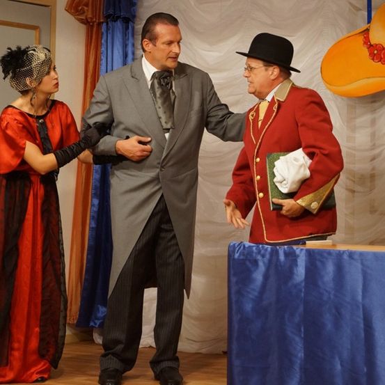 Drei Personen auf einer Bühne, eine Frau in Rot und Schwarz, ein Mann in einem grauen Anzug und ein Mann in einem roten Mantel mit Hut.