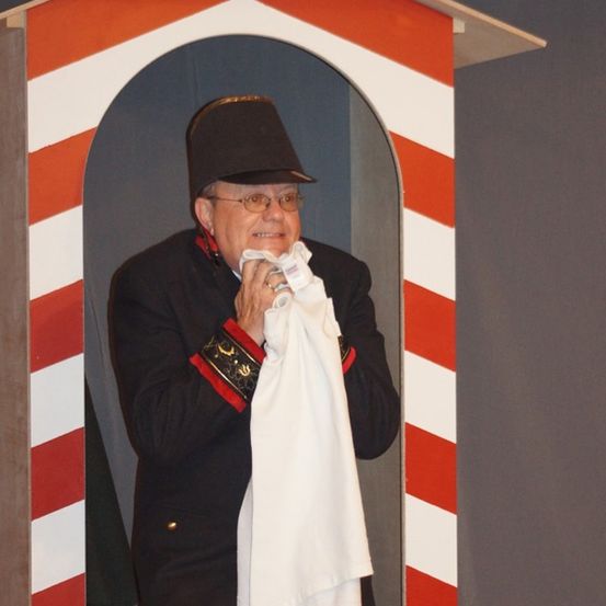 Ein Mann in einem schwarzen Uniform steht in einer rot-weiß gestreiften Kabine und hält ein weißes Tuch. Er scheint zu lächeln.