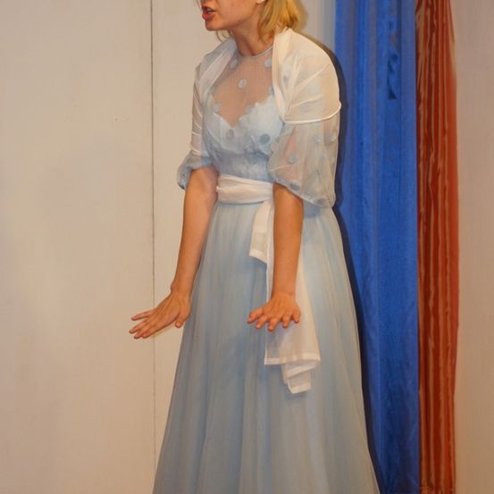 Eine Frau in einem hellblauen Kleid mit einem weißen Umhang steht vor einer weißen Wand, trägt eine Brille und spricht. Hinter ihr befinden sich blaue und orangefarbene Vorhänge.