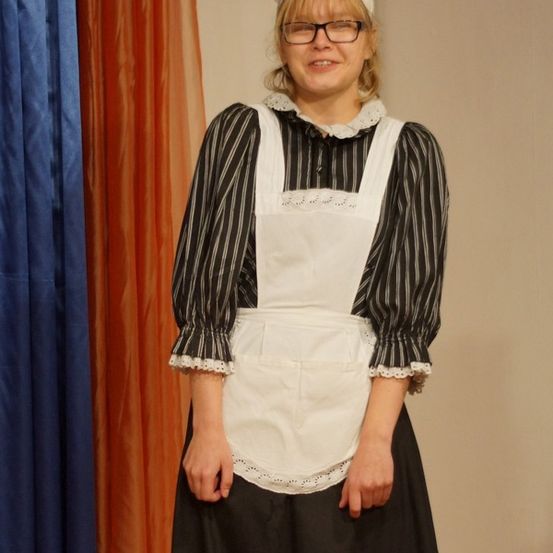 Eine Frau mit Brille, die eine weiße Schürze und ein schwarz-weißes Oberteil trägt, steht lächelnd vor einem blauen und orangefarbenen Vorhang.