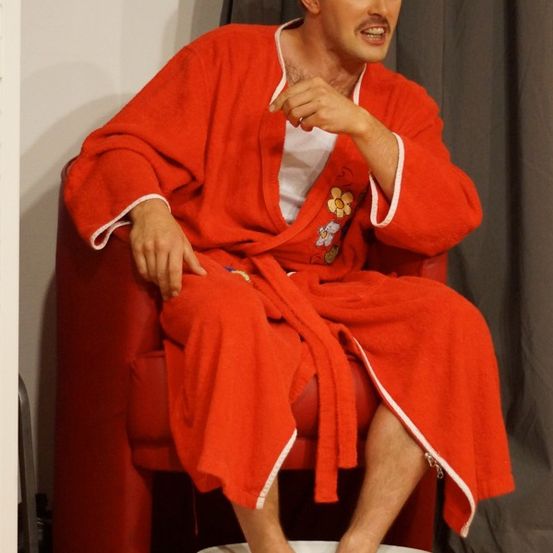 Ein Mann in einem roten Bademantel sitzt auf einem roten Stuhl mit den Füßen in einer weißen Schüssel und lächelt und redet.